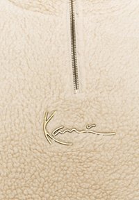 Beige fleece genser med halv glidelås, med en teksturert overflate og en gyllen skrifttype-logo på forsiden. Mykt stoff og enkelt design.