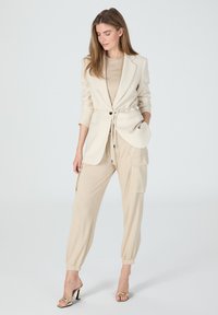 Beige tailored blazer with tie waist, layered over a light beige top, paired with light beige joggers featuring side pockets and elastic cuffs.