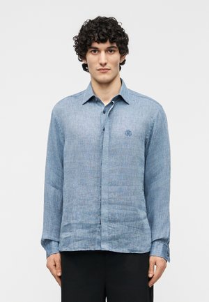 Homme aux cheveux bouclés portant une chemise en lin bleu clair à manches longues avec des poignets à boutons et un pantalon noir, debout devant un fond uni.