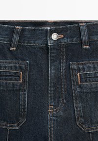 Dark blue denim jeans featuring bronze stitching, a metal button closure, and two front pockets with stitched detailing.