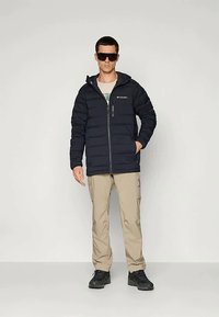 Ναυτικό puffer μπουφάν με φερμουάρ, κρουστό σχέδιο και κουκούλα, συνδυασμένο με μπεζ cargo παντελόνι και μαύρα παπούτσια, μοντέλο που φοράει σκούρα γυαλιά ηλίου.