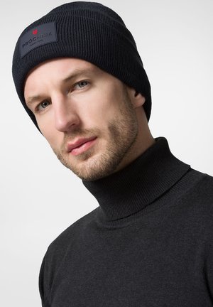 Czarna, prążkowana czapka beanie z naszywką logo, noszona z czarnym golfem. Tkanina wydaje się miękka i przytulna, akcentując neutralną paletę kolorów.