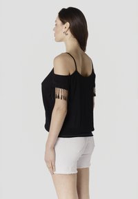 Mujer de perfil vistiendo una blusa negra con hombros descubiertos y flecos, y pantalones cortos de mezclilla blancos, sobre un fondo liso y claro.