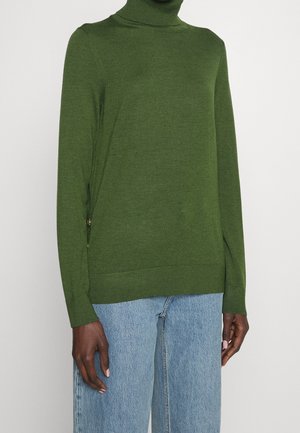 Osoba v zeleném roláku a světlých modrých džínách stojící před jednobarevným bílým pozadím.