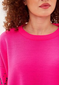 Ljusrosa tröja med ribbad halsringning och strukturerad stickning. Designen inkluderar subtila gröna detaljer på ärmarna.