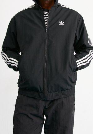 Homme portant une veste de survêtement Adidas noire avec des bandes blanches sur les manches, fermeture éclair devant et logo Adidas sur la poitrine, mains dans les poches.