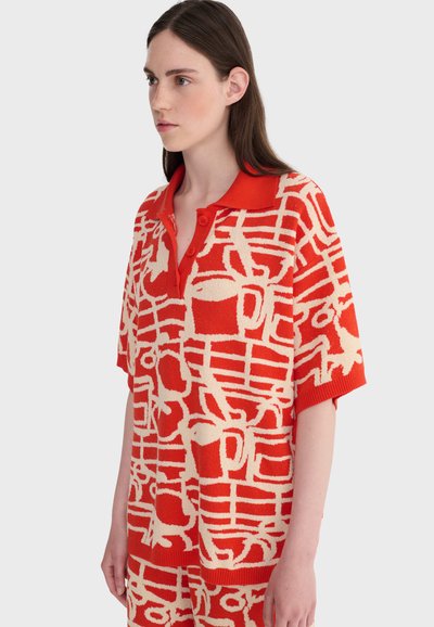 Jeune femme portant un polo en tricot à motifs rouges et beiges à manches courtes, debout devant un fond clair uni.