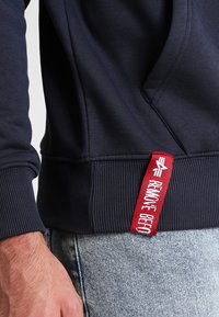 Marineblauwe sweatshirt met ribbelboorden en -zoom. Aan de zijkant bevindt zich een rode tag met de tekst "VERWIJDER VOOR", wat een contrasterend detail biedt.