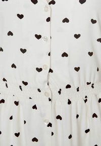 Robe blanche ornée d'un imprimé de petits cœurs noirs éparpillés, avec un devant boutonné et une taille smockée. Le tissu a une texture douce et un léger éclat.