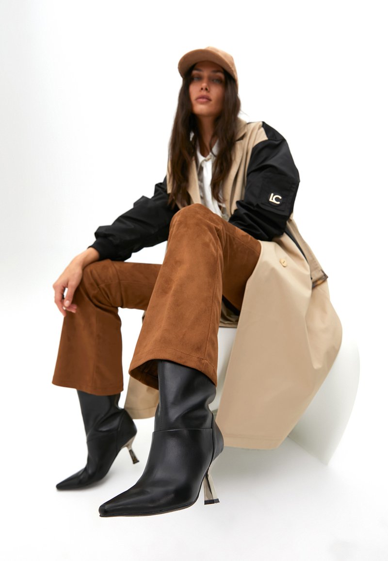 Czarne skórzane botki za kostkę z ostrymi noskami i metalowymi obcasami, zestawione z brązowymi zamszowymi spodniami o szerokich nogawkach oraz beżową i czarną kurtką.