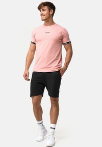 Tricou din bumbac roz deschis cu detaliu logo negru pe piept, asortat cu pantaloni scurți negri și adidași albi, având șosete albe cu dungi.