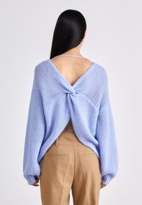 Kvinna med långt svart hår som bär en ljusblå stickad tröja med öppen rygg och beige högmidjade byxor, vänd bort.