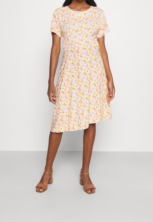 Femme portant une robe rose longueur genou avec un motif floral jaune et blanc et des manches courtes, associée à des sandales à talons blocs ouvertes couleur beige.
