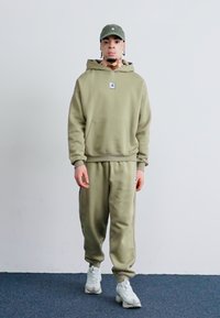 Olivengrøn hættetrøje og matchende joggers lavet af blødt stof, med et logo-patch, ribbede manchetter og hvide sneakers.