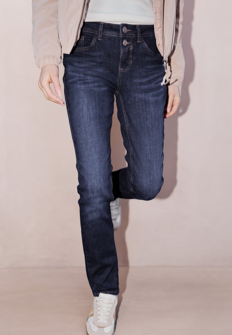 Personne portant un jean slim bleu foncé, une veste beige clair, un haut blanc et des baskets blanches, marchant devant un fond clair uni.