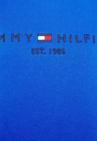 Camiseta de algodón azul con "TOMMY HILFIGER" bordado en hilo oscuro, junto a un pequeño diseño de bandera y "EST. 1985" debajo.