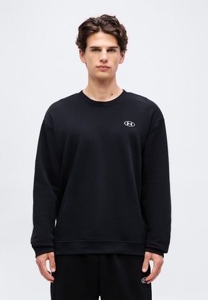 Young man with short dark hair wearing black Under Armour sweatshirt and matching pants, standing against plain light background.