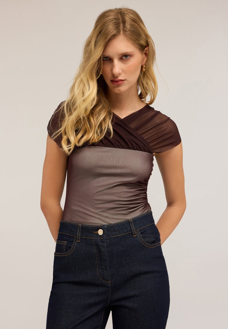 Donna con lunghi capelli biondi che indossa una maglietta corta trasparente marrone e jeans blu scuro, in piedi con le mani dietro la schiena.