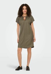 Olivenfarbenes Kleid mit V-Ausschnitt und Bindedetail, kurzen Ärmeln, knielanger Schnitt, aus glattem Stoff. Mit schwarzen Schuhen getragen.