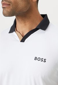 Weißes Poloshirt mit strukturiertem Muster, schwarzem Kragen und einem braunen Akzent unterhalb des Kragens. "BOSS"-Logo auf der Vorderseite.