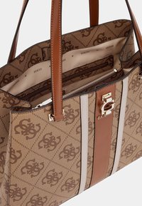 Sac fourre-tout marron à motifs avec une fermeture éclair brillante, des accents en cuir crème et brun clair, ainsi qu'un détail de matériel logo doré.