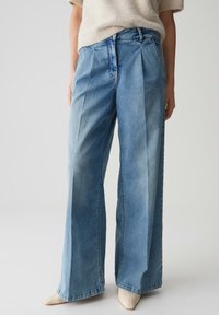 Hoge taille, wijde blauwe jeans met plooien aan de voorkant, gedragen met een romige fuzzy trui met korte mouwen en beige puntige schoenen.