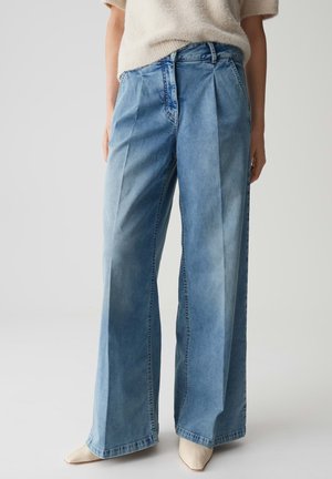 Jeans bleu taille haute à jambes larges avec plis sur le devant, portés avec un pull crémeux en peluche à manches courtes et des chaussures beiges à bout pointu.