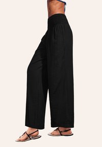 Zwarte wijde broek van lichtgewicht stof, met een smocktail. Vloeiende ontwerp. Draagt met platte sandalen.