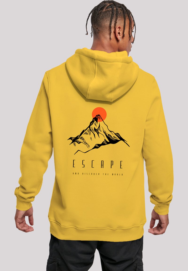 Man wearing yellow hoodie with mountain and red sun graphic and the word "ESCAPE" on the back, standing against plain background.