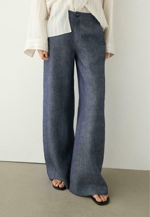 Persona che indossa pantaloni di jeans blu a vita alta e gamba larga con una cintura con bottoni, abbinati a sandali neri aperti e una camicetta bianca dalla trama strutturata.