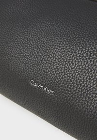 Portefeuille en faux cuir noir avec une surface texturée et le logo "Calvin Klein" en argent sur le devant. Fermeture zippée sur le dessus.