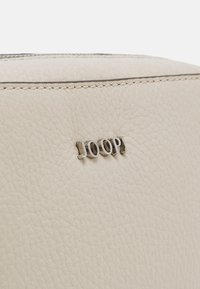 Béžová kožená taška s texturovaným povrchem, na přední straně s logem "JOOP!" ve stříbrné barvě, dokončena pevným prošíváním a zipovým uzávěrem.