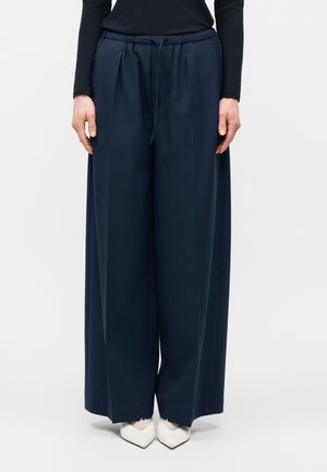 Nő széles szárú, sötétkék, húzózsinóros nadrágban és fehér, hegyes orrú cipőben, egy egyszínű háttér előtt áll.