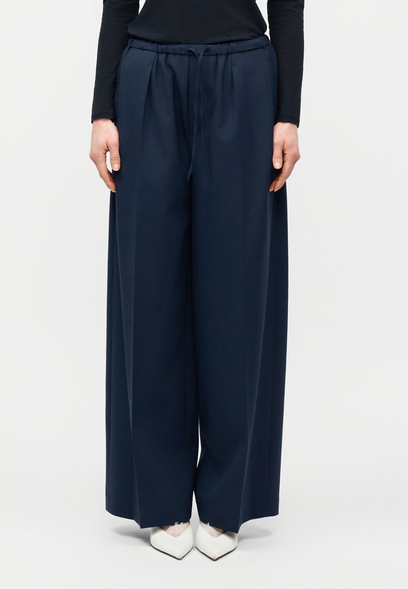 Γυναίκα που φοράει ναυτικό μπλε παντελόνι με φαρδιές παντελόνες και ζωνάρι στη μέση, καθώς και λευκά παπούτσια με μυτερή μύτη, στέκεται μπροστά σε απλό φόντο.