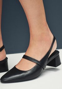 Zapatos slingback de cuero negro con punta afilada, una cinta ancha a lo largo del empeine y un tacón bajo en bloque. Textura suave con mínimos acentos.
