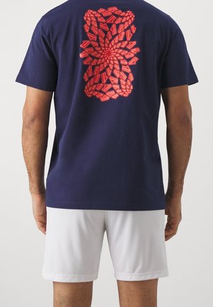 Mørkeblå bomulds t-shirt med et livligt rødt svungent grafisk mønster på ryggen. Korte ærmer og afslappet pasform. Hvide shorts synlige.
