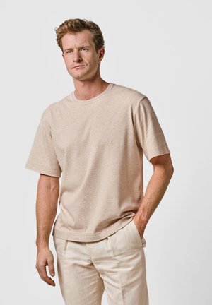 Hombre con cabello rizado castaño claro, vestido con una camiseta de manga corta beige y pantalones claros, de pie con una mano en el bolsillo.