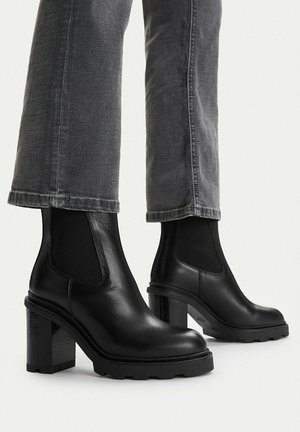 Stivaletti alla caviglia con tacco in pelle nera e pannelli laterali elastici, indossati con jeans grigi in denim.