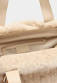 Sac à main beige avec une doublure en fausse fourrure douce et des accents en cuir. Comprend une fermeture éclair et une poche intérieure avec une étiquette de logo.