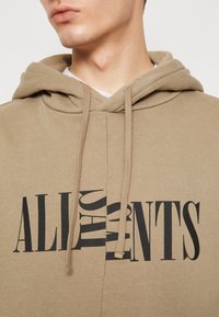 Beżowa bluza z kapturem z przodu z dużym, pogrubionym napisem "ALL" w czarnych literach. Posiada regulowany sznurek przy kapturze i miękką fakturę.
