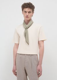 Camicia beige a maniche corte indossata con una sciarpa verde legata al collo, abbinata a pantaloni strutturati marrone chiaro. Design minimalista, tessuto morbido.