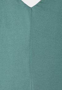 Top verde menta con scollatura a V realizzato in un tessuto liscio e testurizzato. Presenta una cucitura centrale che scorre verticalmente lungo la schiena.