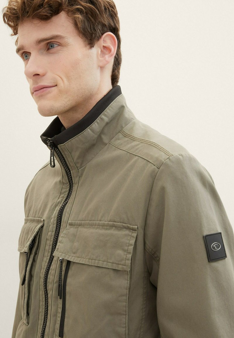 Chaqueta verde oliva con alto cuello, frontal con cremallera y dos bolsillos en el pecho; presenta detalles en negro y un parche de logo en la manga.