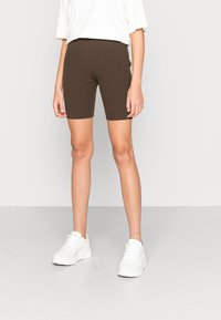 Persona vistiendo pantalones cortos de punto acanalado marrones, camiseta blanca de gran tamaño y zapatillas gruesas blancas, sobre un fondo llano de suelo gris y pared blanca.