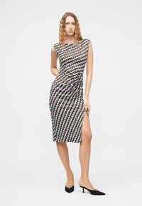 Γυναίκα με μακριά κυματιστά μαλλιά, ντυμένη με αμάνικο midi φόρεμα με σχέδια και πλευρική σχισμή, φορώντας μαύρα παπούτσια με χαμηλό τακούνι, στέκεται μπροστά σε λευκό φόντο.