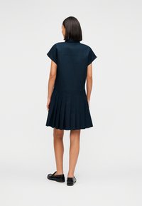 Μπλε σκούρο φόρεμα με κοντά μανίκια, πλισέ φούστα και γιακά. Διαθέτει απλό ύφασμα και εφαρμοστή μέση. Μαύρα παπούτσια χωρίς κορδόνια ολοκληρώνουν το σύνολο.