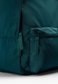 Coin d'un sac en nylon vert foncé avec deux poches zippées, chacune équipée d'une languette en tissu avec un détail discret en texte.