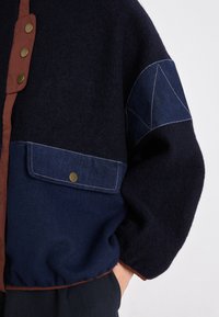 Giacca in misto lana blu navy con patta frontale con bottoni marroni, dettagli in denim e tasca frontale con finiture in ottone. Finitura testurizzata.