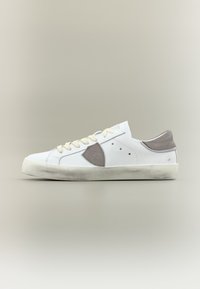 Baskets basses blanches avec lacets beige, talon en daim gris et patch logo, et semelle en caoutchouc légèrement usée sur un fond neutre.