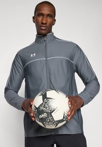 Gray athletic jacket with white accents, high collar, and full zipper. The model holds a black and white patterned soccer ball.